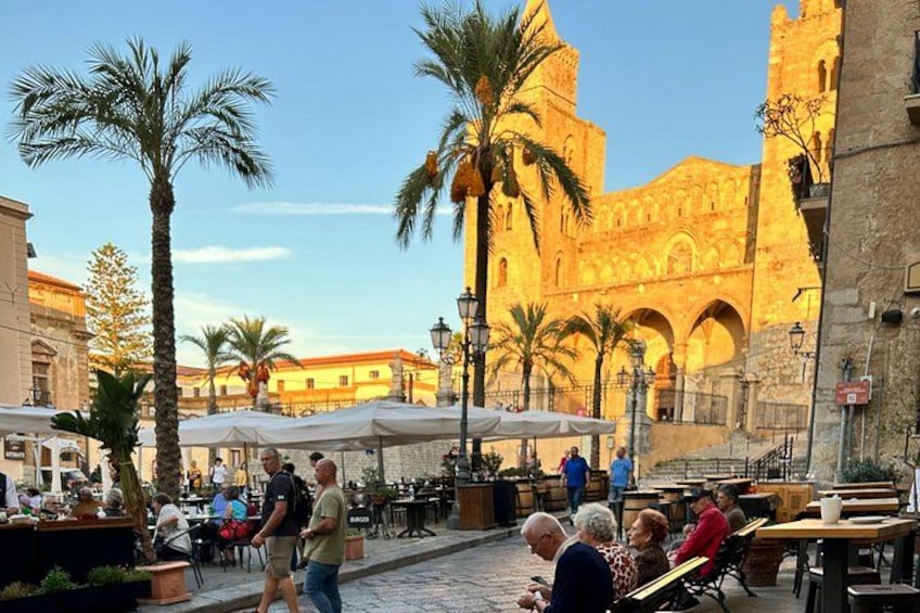 A plaza with palm trees, outdoor seating, and a historic building with arches in the background. 1