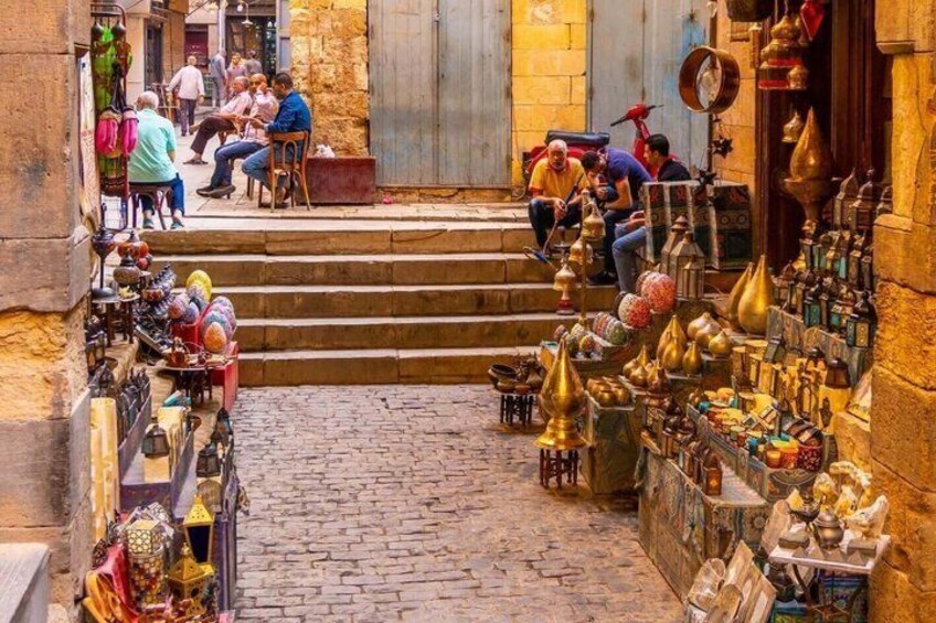 Une ruelle étroite bordée de boutiques de chaque côté, des marches menant à un immeuble, et des gens assis et debout. 3