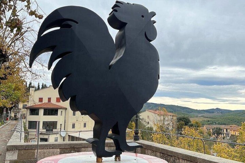 En stor, svart skulptur föreställande en tupp, placerad på en piedestal, med en vacker utsikt över byggnader och kullar i bakgrunden. 3