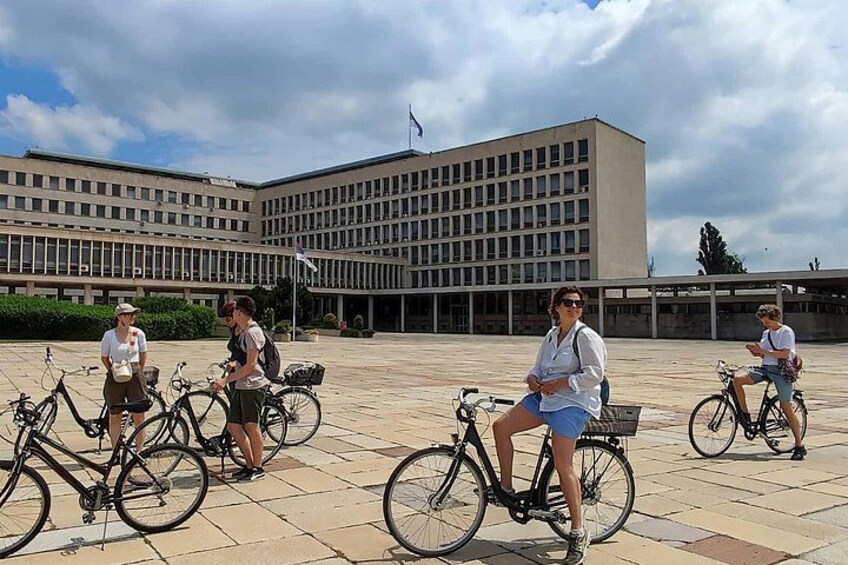Four people on bicycles in front of a large building with a flag. 5
