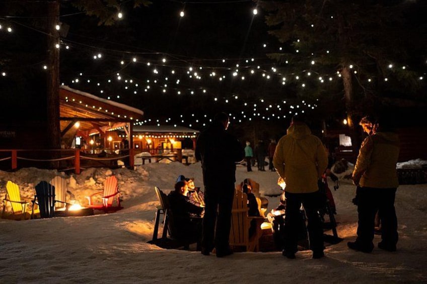 Een besneeuwde buitenruimte met lichtjes, een houten constructie en mensen rond een vuur. 2