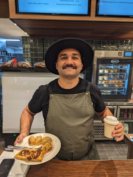 A person in a black shirt and green apron holding a plate of food and a coffee cup. 3