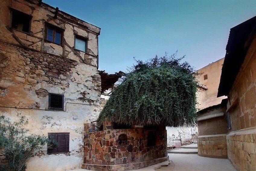 A narrow alley with stone buildings, a tree with a thatched roof, and a clear blue sky. 4