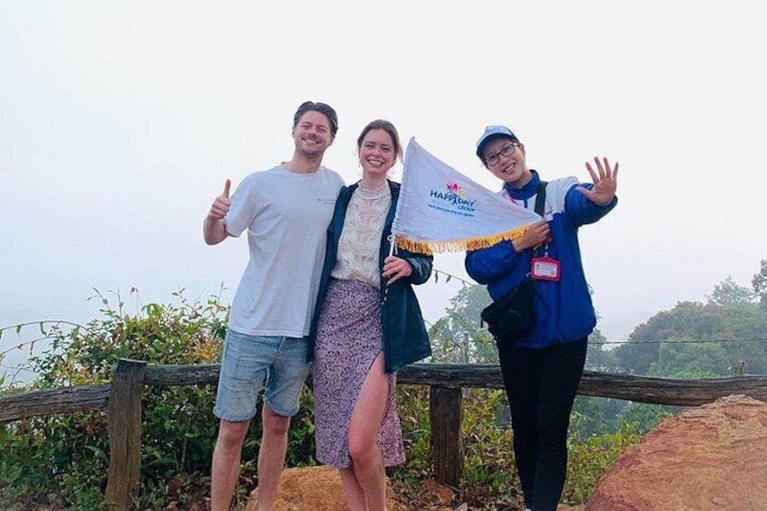 Three people standing on a wooden platform with a flag, surrounded by greenery and hills. 4