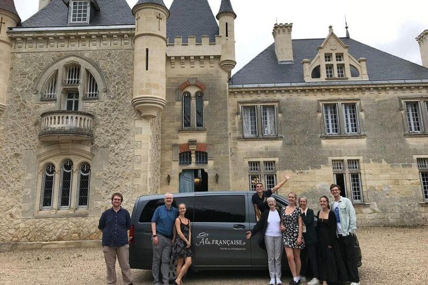 A group of people standing in front of a large stone building with multiple windows and a parked vehicle. 2