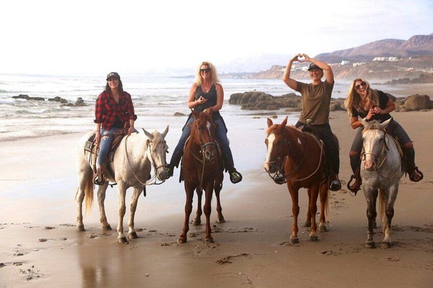 Quatre personnes à cheval sur une plage, avec l’océan et des collines rocheuses en arrière-plan. 4