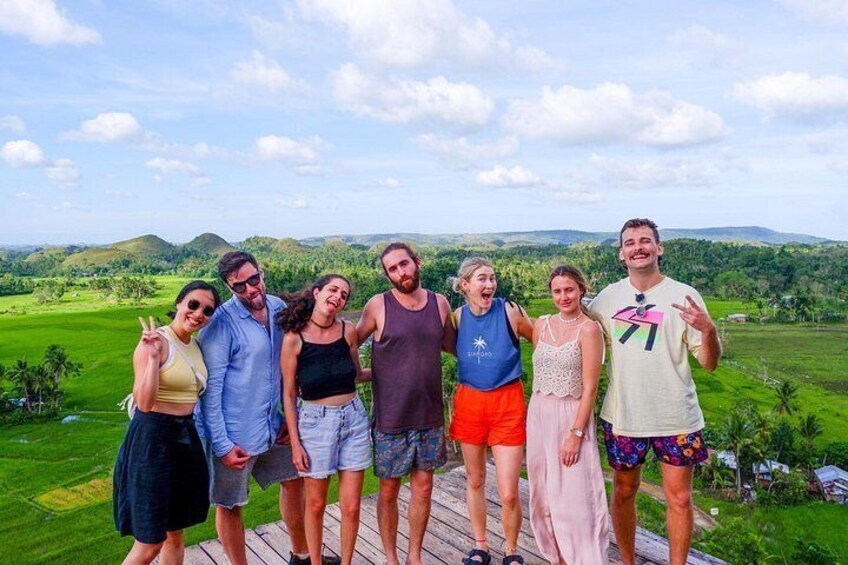 A group of people standing on a wooden deck with a scenic view of green hills and a clear sky. 1