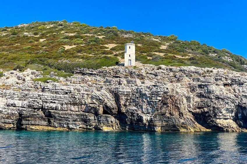 Un acantilado costero con un faro en la cima, agua azul cristalina y un paisaje montañoso con vegetación escasa. 1