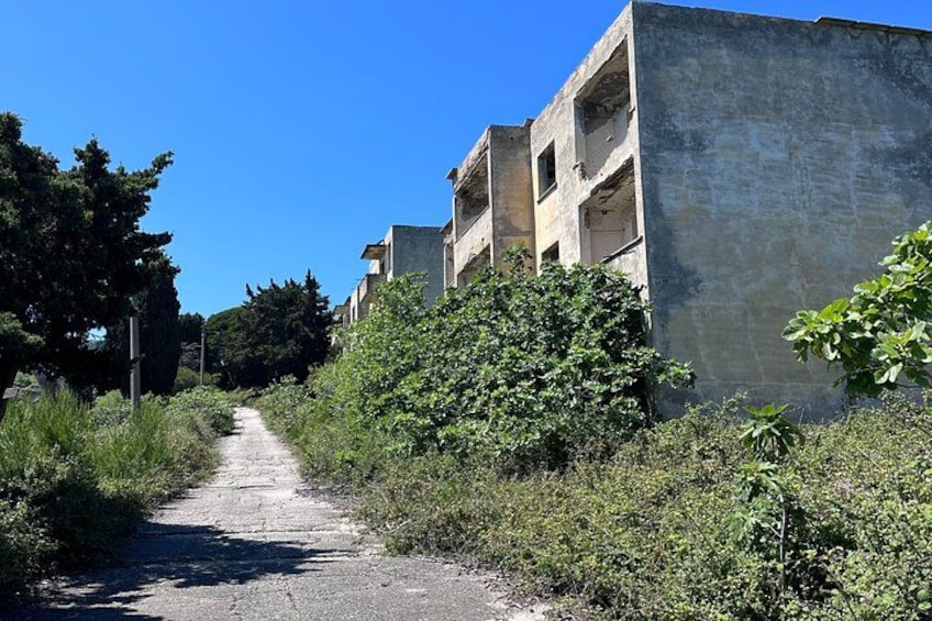 Ein schmaler, unbefestigter Weg führt zu einer Reihe verlassener Gebäude mit überwucherter Vegetation. 5