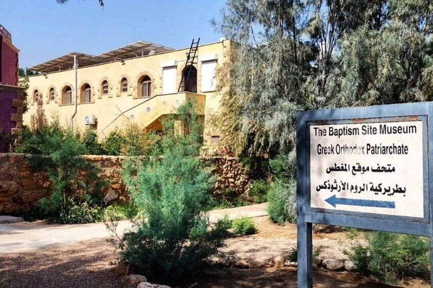 A sign for The Baptism Site Museum Greek Orthodox Patriarchate with a building in the background. 2
