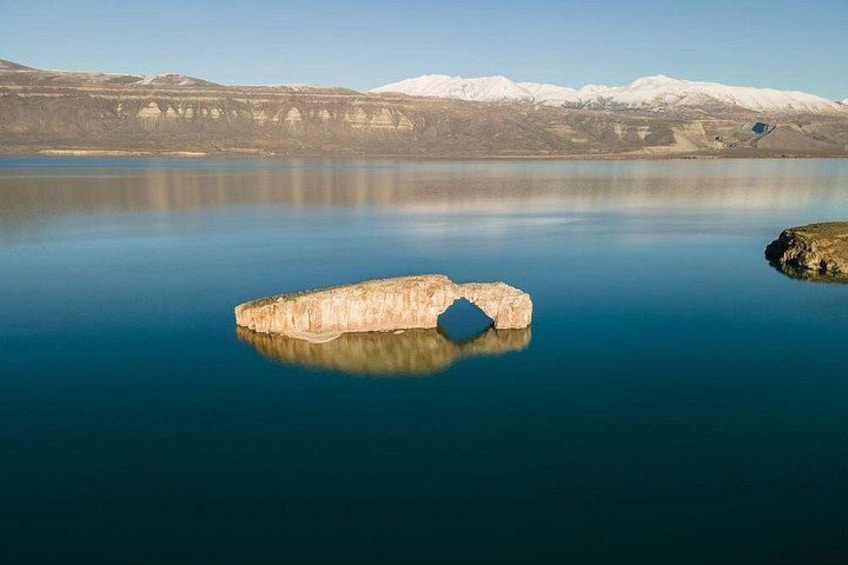 A calm lake with a large rock formation in the center and snow-capped mountains in the background. 1