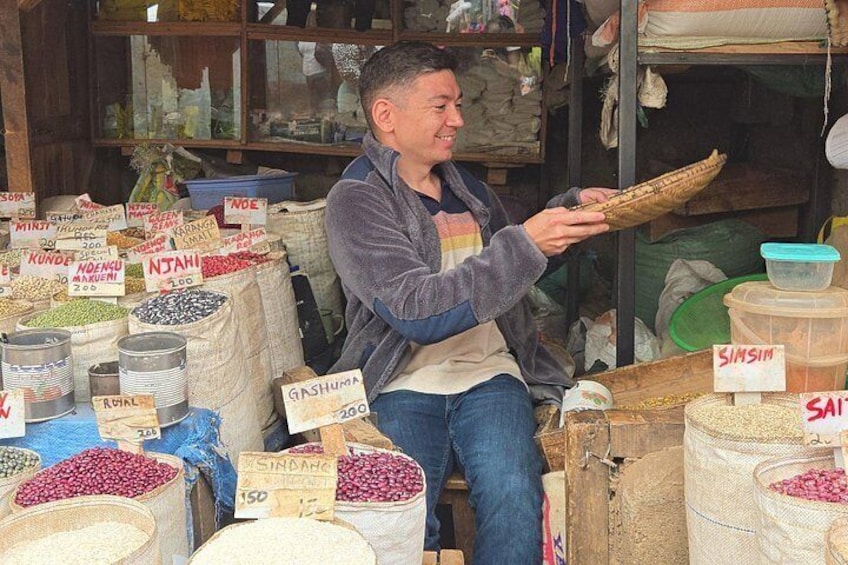 Une personne est assise derrière un étal de marché, devant différents sacs contenant des céréales et des légumineuses, dont certains sont étiquetés avec les prix. 3