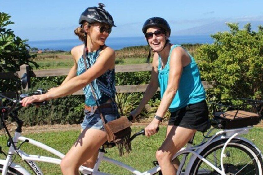 Two people riding bicycles on a grassy area with a wooden fence and greenery in the background. 3