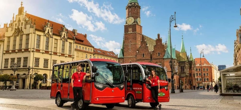 Zwei rote Busse stehen auf einem historischen Platz mit einem markanten Glockenturm und verzierten Gebäuden im Hintergrund. 1