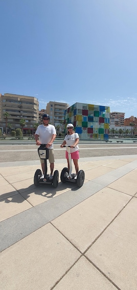 Kaksi ihmistä, mies ja nainen, ajelevat segway-lautoilla kaupunkimaisemassa, jossa on moderneja rakennuksia ja kirkas taivas. 2
