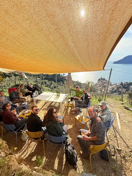 Un grupo de personas sentadas en una mesa al aire libre bajo una toldilla, con vistas a un paisaje costero con colinas y una masa de agua. 4