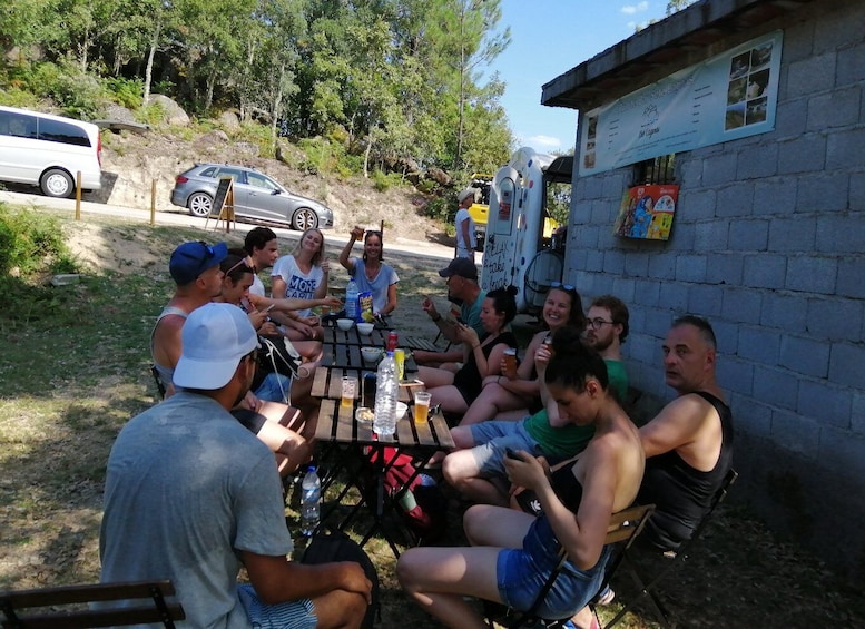 Un groupe de personnes est assis autour d’une table en bois à l’extérieur, avec un bâtiment et des voitures garées en arrière-plan. 2
