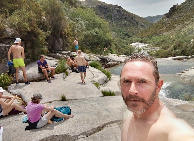 A group of people sitting and standing on rocks by a river surrounded by greenery and hills. 5