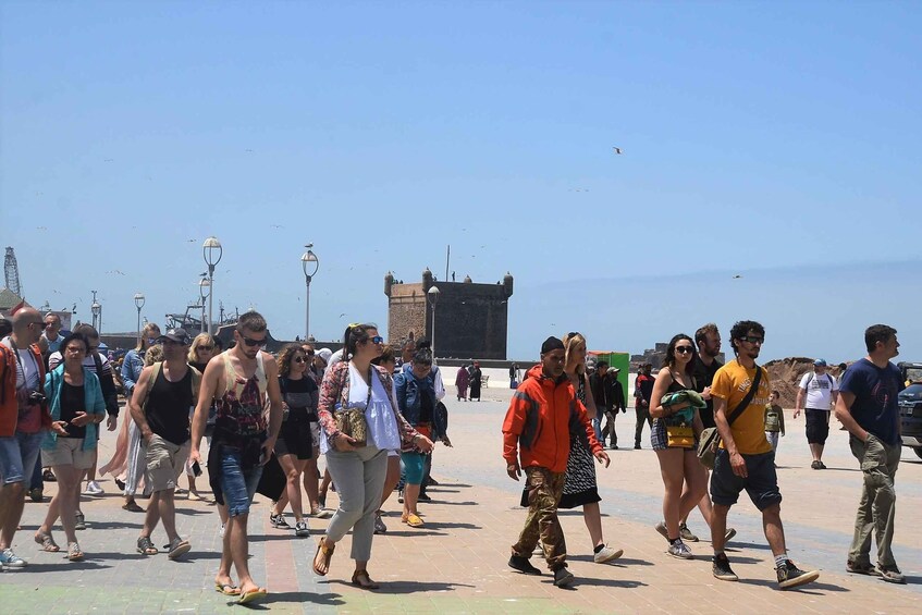 A crowded promenade with people walking, a large historical building in the background, and a clear blue sky. 3