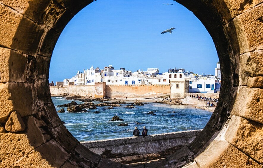A coastal town with white buildings, a stone archway, and the sea. 1