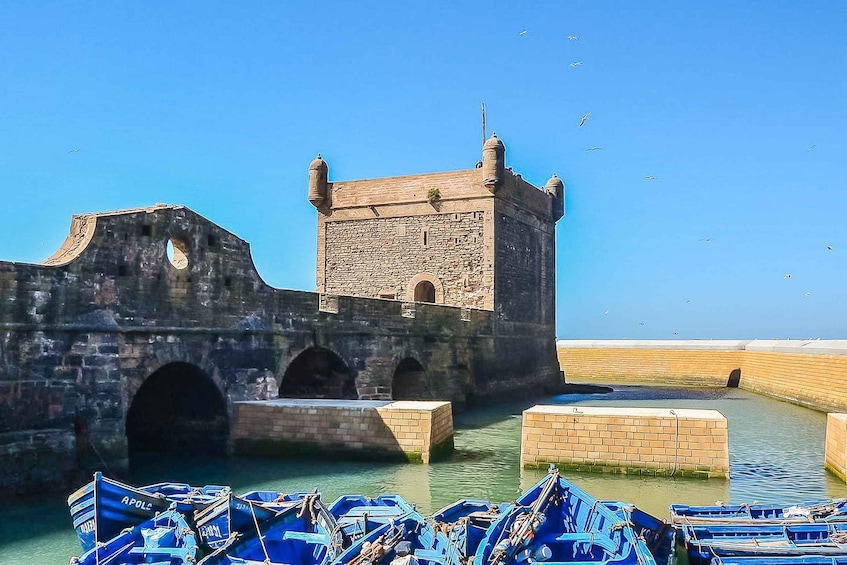 A historic stone tower with arched gateways, surrounded by calm water and small boats. 1