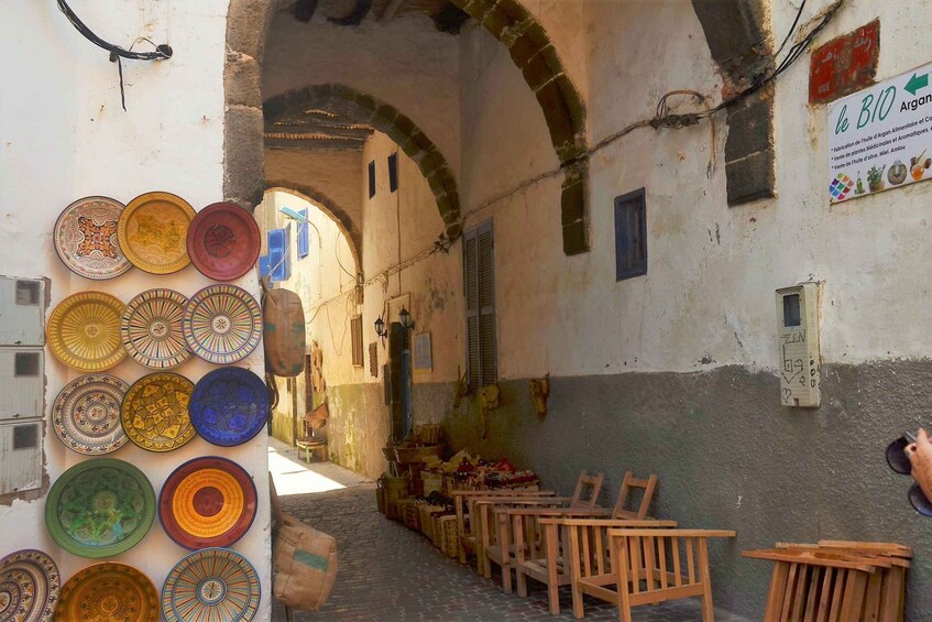 A narrow alleyway with colorful plates on a wall, wooden chairs, and a sign indicating a local market. 5