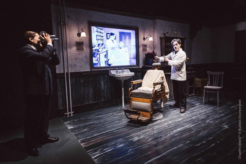 A man in a white lab coat standing next to a vintage barber chair, with a television screen displaying a scene in the background. 4