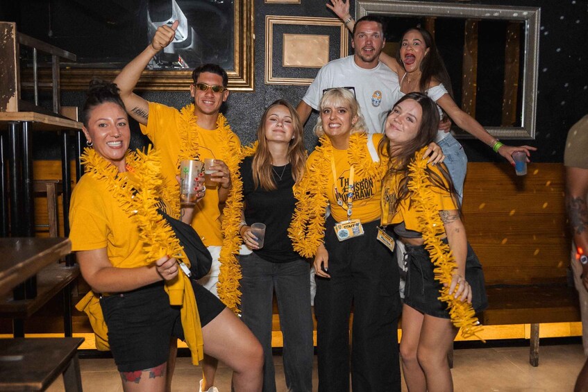 A group of people wearing yellow shirts and leis, gathered in a room with a wooden floor and a black wall with framed pictures. 3