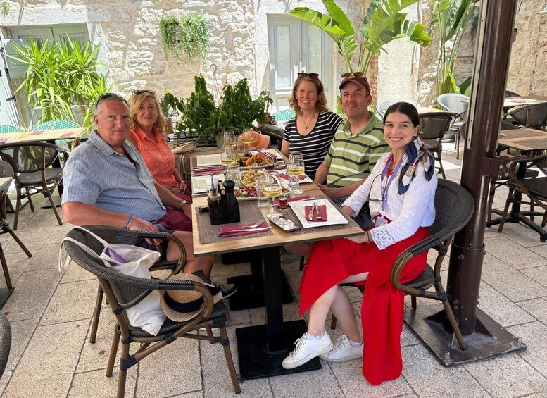 A group of people seated at an outdoor table with food and drinks, surrounded by chairs and plants. 3
