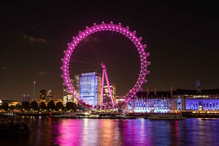 A large Ferris wheel with pink lights, a river, and buildings in the background. 2