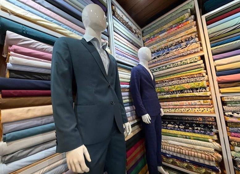 Two mannequins in suits, one in a dark suit and the other in a blue suit, standing in front of a wall of folded fabrics in various colors and patterns. 4