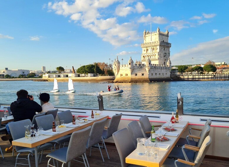 En restaurant med borde med udsigt over en flod, et slot i baggrunden og sejlbåde på vandet. 1