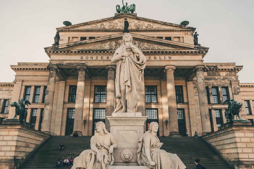 A grand neoclassical building with a central statue and two seated figures on its facade. 4