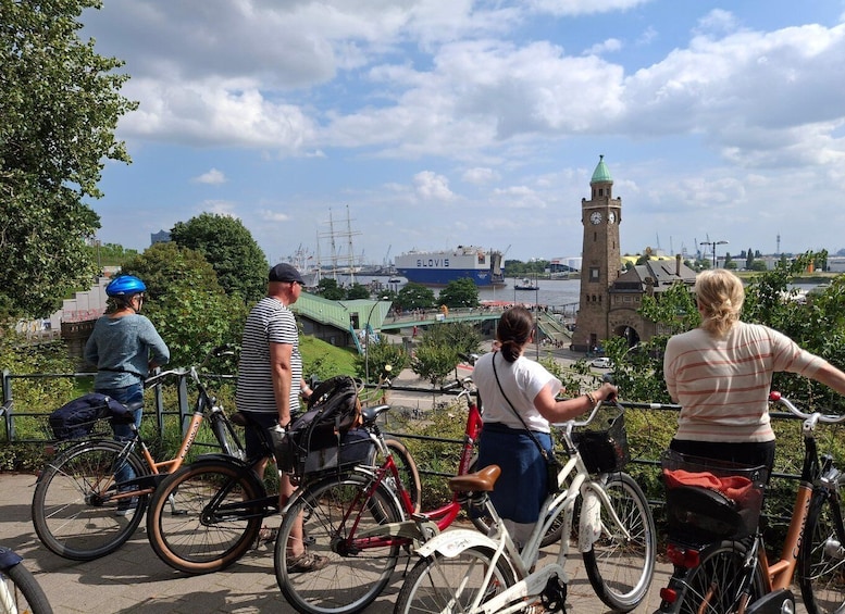 Un groupe de cyclistes se trouve sur un pont surplombant une rivière, avec une tour de l’horloge historique et un grand navire en arrière-plan. 1