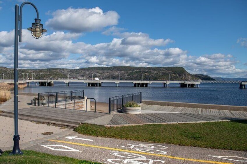 A waterfront promenade with a wooden boardwalk, metal railings, and a bridge in the background. 3
