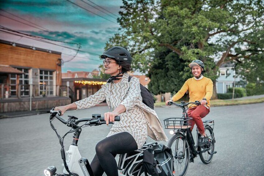 Two people riding bicycles on a street with buildings and trees in the background. 1