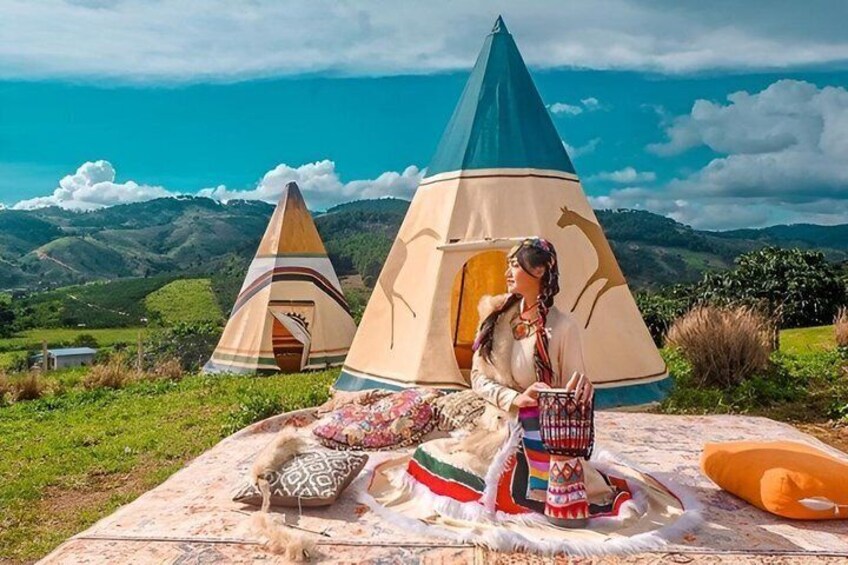 A person sitting on a blanket with a colorful blanket and pillows, near a conical tent in a grassy area with hills in the background. 2