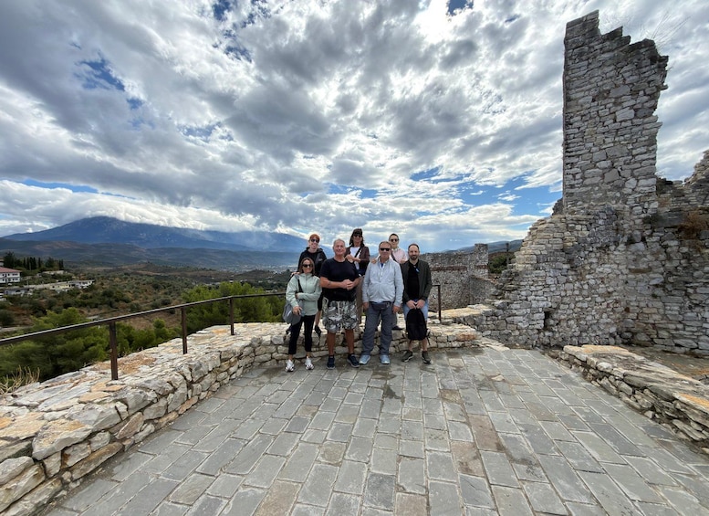 A group of people standing on a stone platform with a stone wall and ruins in the background, overlooking a mountainous landscape. 4