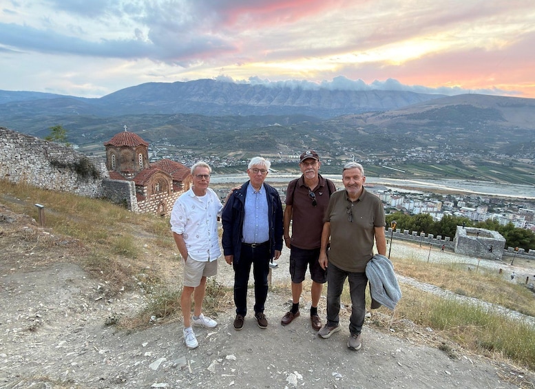 Four people standing on a rocky path with a view of a mountainous landscape and a church in the background. 2