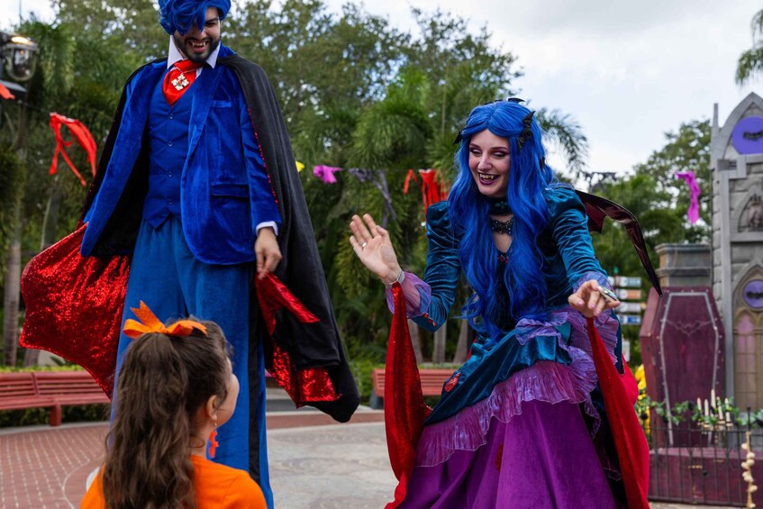 A person in a blue costume with a red sash and a black cape, holding a red object, standing next to a person in a blue and purple costume with a black cape, in an outdoor setting with trees and a building in the background. 2