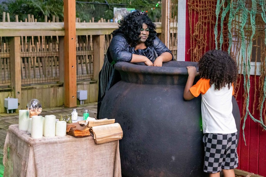 A person in a black and white outfit leaning on a large black barrel, a child in an orange shirt and checkered shorts standing beside it, and various items like candles and a crystal ball on a cloth-covered table. 3