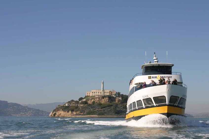 A boat named Blue Gold Fleet moving through the water with a lighthouse and a building on a cliff in the background. 1