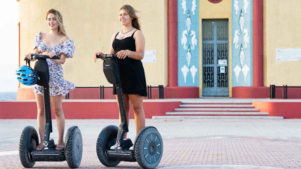 Two people riding segways on a paved area with a building in the background. 2