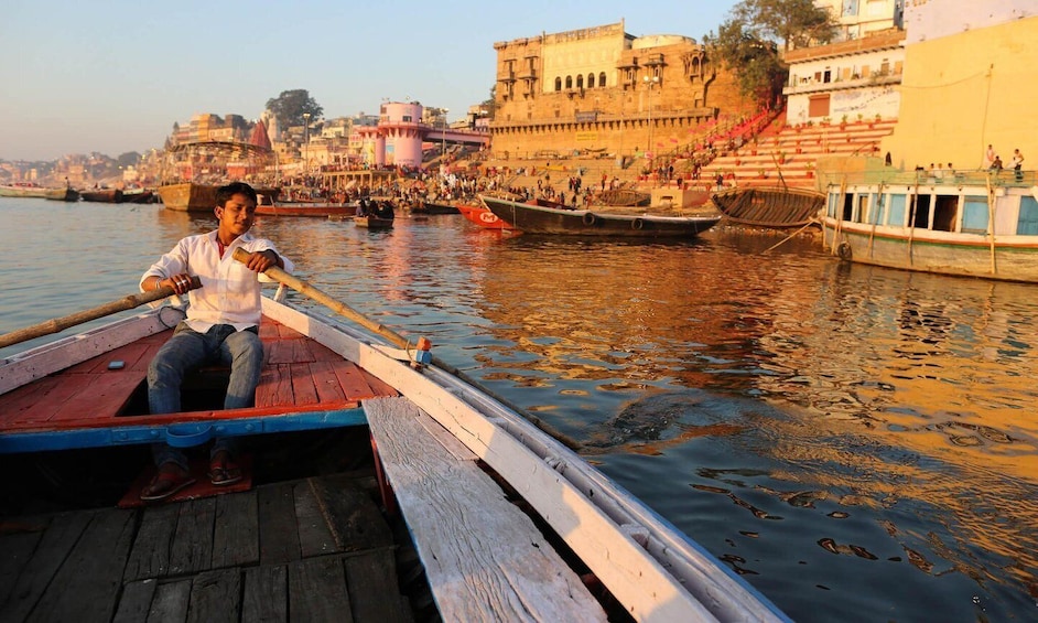 A man rowing a wooden boat on a river with buildings and steps in the background. 3