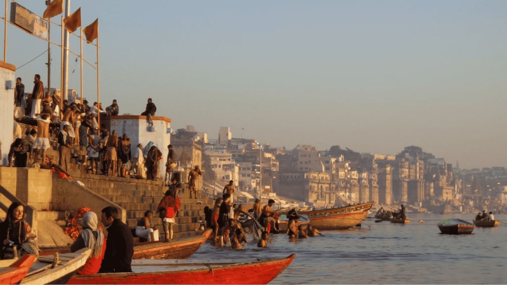 A crowded riverside with people in boats and on the steps, and a cityscape in the background. 2