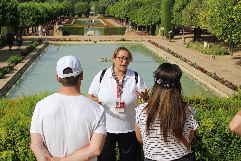 Eine Gruppe von Menschen steht an einem Teich in einem Park, im Hintergrund sind Bäume und eine Statue zu sehen. 1