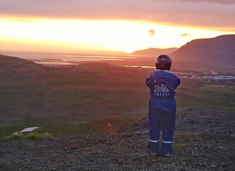 A person in a blue jumpsuit with a helmet, standing on a rocky terrain, overlooking a valley with a sunset in the background. 5