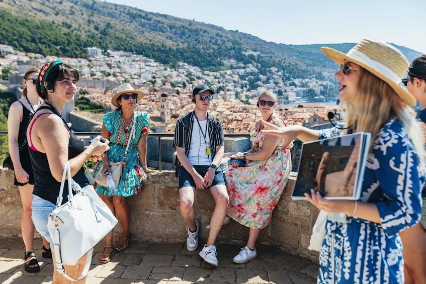 A group of people sitting on a stone wall overlooking a coastal town with buildings and hills in the background. 2