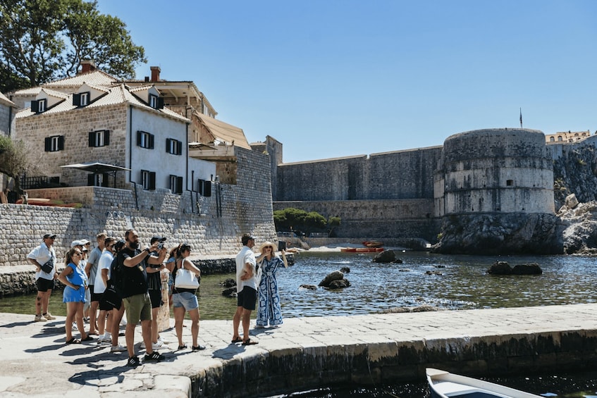 A group of people standing by a waterfront with a historic stone building and a large fort in the background. 4