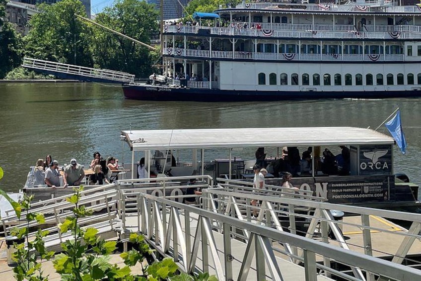 A river with a large white boat docked at a pier, a smaller boat with people on it, and a bridge in the background. 3